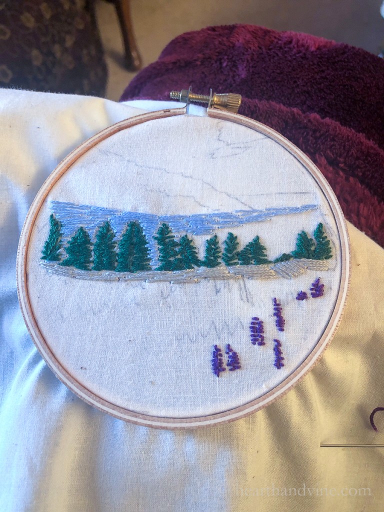 Embroidery hoop with a scene of partially embroidered evergreen trees and purple flowers.