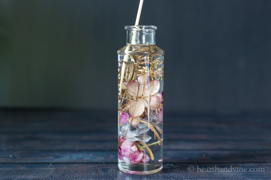 Dried flowers in oil in a tall glass jar with a wooden skewer used to move flowers around.