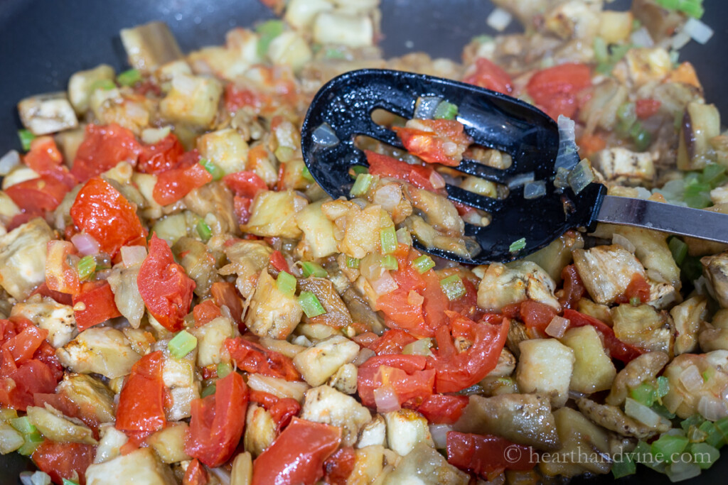 Sauted eggplant tomatoes, onions, celery and other items. A plastic slotted spoon pushes down on vegetables.