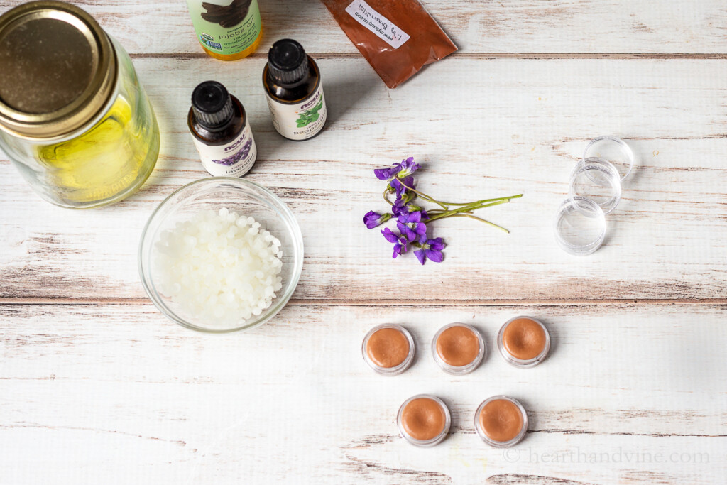 Beeswax pellets, fresh violets, essential oils, mica powder and small containers filled with violet balm/salve.