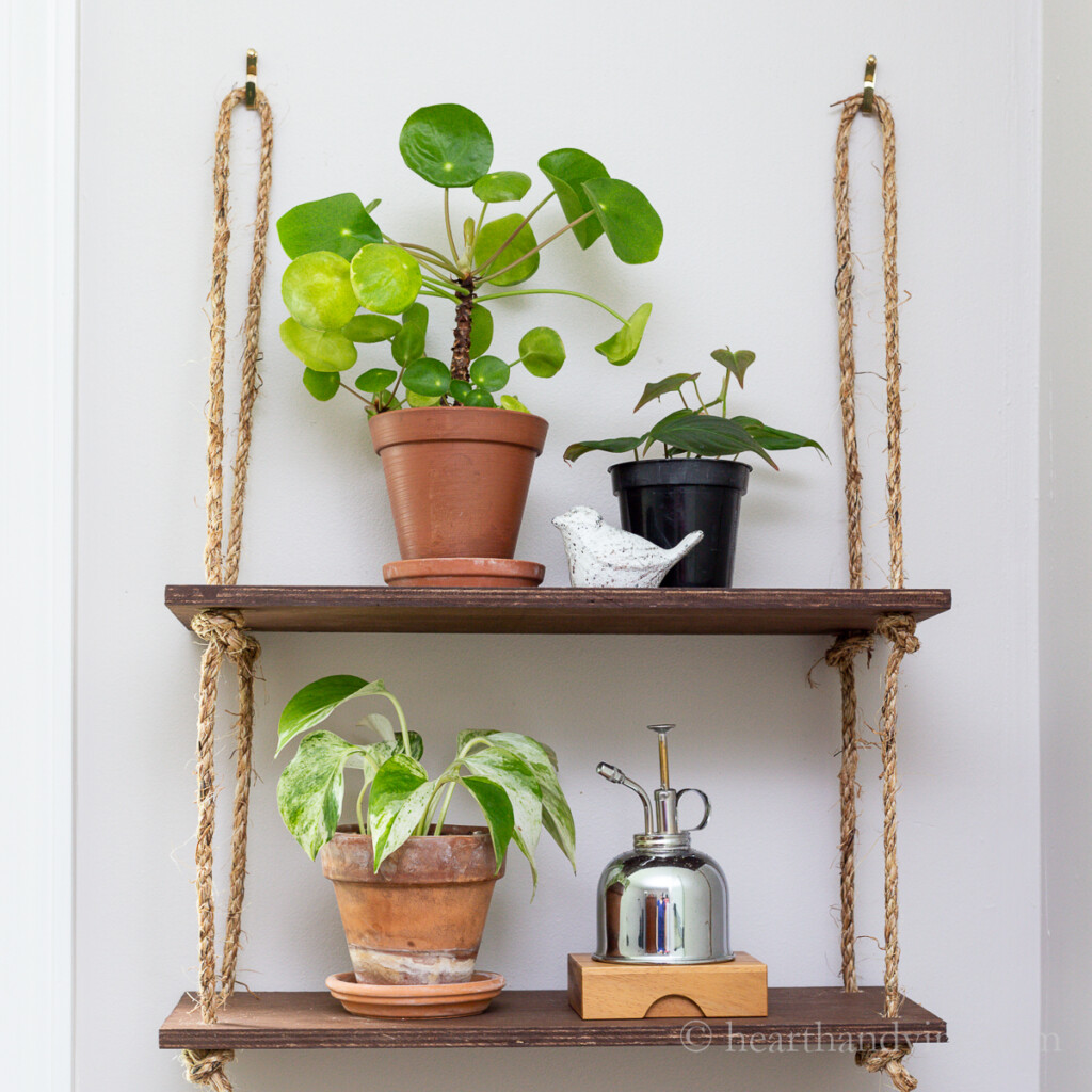 DIY wood hanging rope shelf. Plants and a ceramic bird and a watering can are on the shelves.