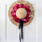 Straw hat wreath with pink and red flowers and a black bow.