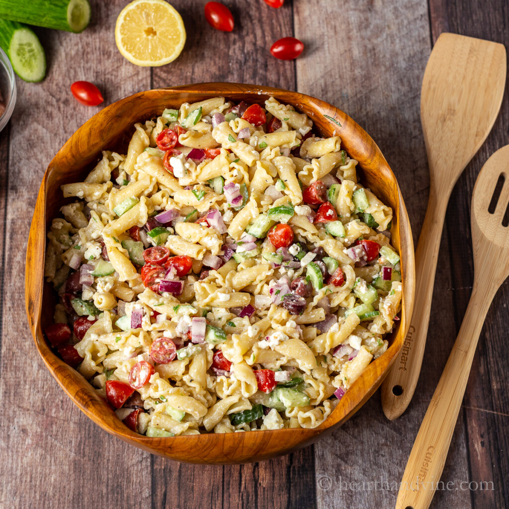 Greek pasta salad in a wooden bowl with two wooden serving utensils, lemon, grape tomatoes and cucumber on the side.