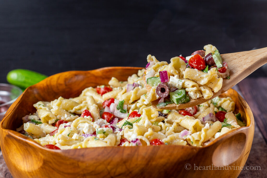 Spoonful of Greek pasta salad coming out of a large wooden serving bowl.