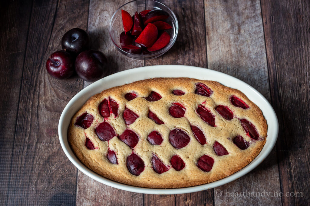 Large oval pan with plum cake next to whole plums and a small bowl of fresh cut plum wedges.