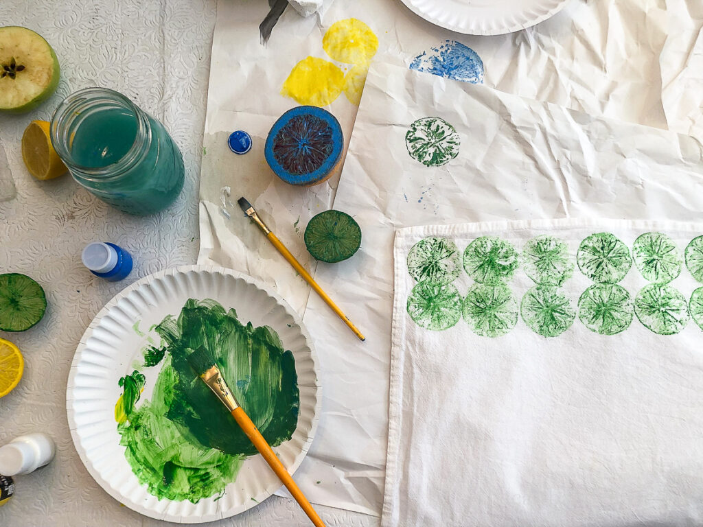 Image showing a workspace with a jar of water, paper plate with paint, a paintbrush and a tea towel with green lime prints.