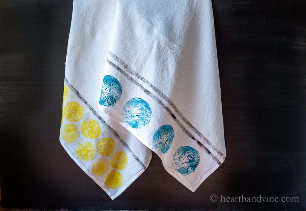 Two fruit hand printed tea towels hanging. One with lemon yellow prints and one with an apple print in blue.