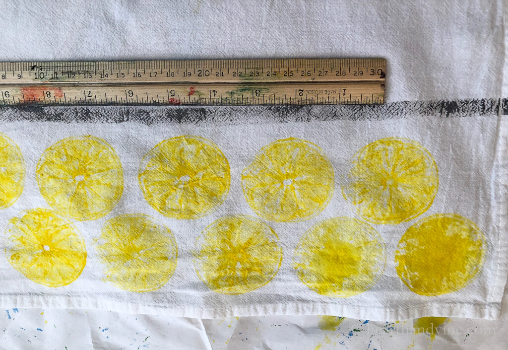 A ruler with a black painted line underneath over a two rows of lemon printed stamp images on a flour sack towel.
