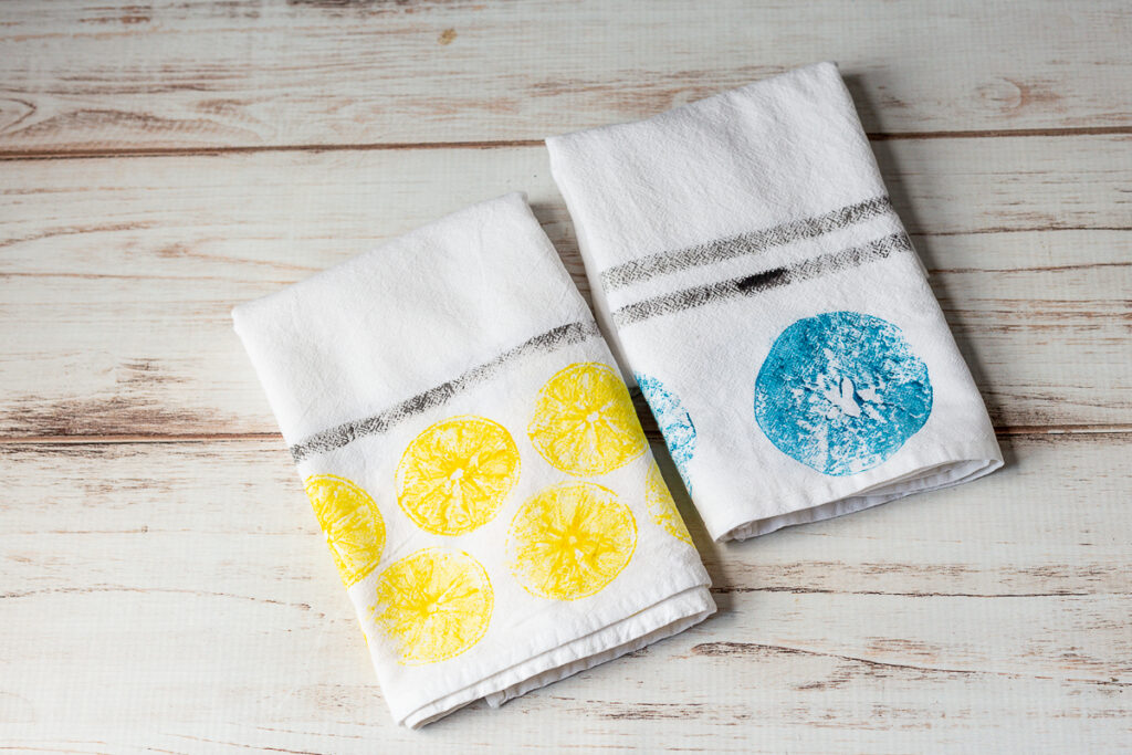 A lemon yellow print and an apple print in blue tea towel with black stripes on  a table.