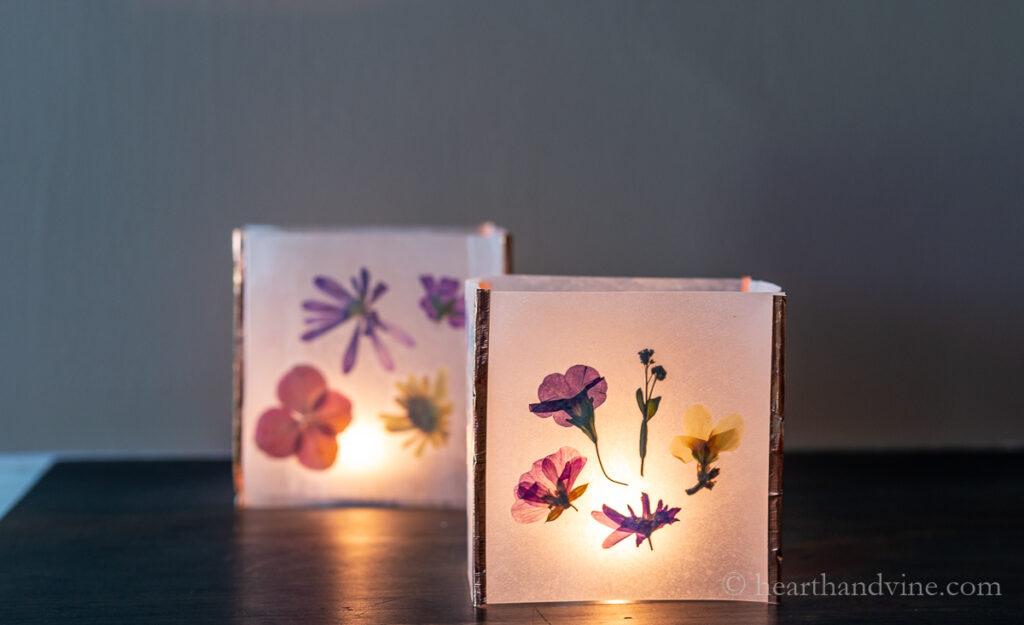 Two copper tape pressed flower luminaries with candles inside in a dark room.