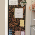 A tall narrow bulletin board with cork backing and black floral stencil on a wall with pinned notes.