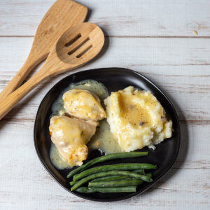 Creamy chicken with gravy, mashed potatoes and green beans next to wooden spoons.