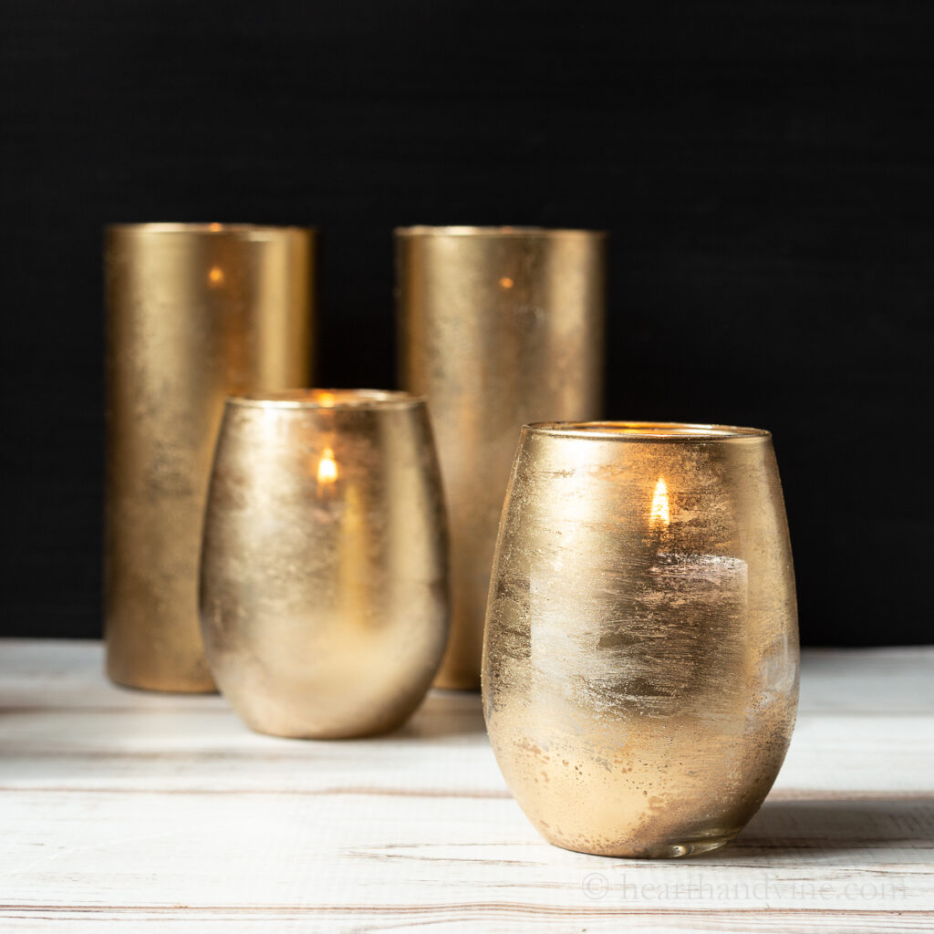 Four gold mercury glasses with candles. Two rounded in front and two taller and  straight in the back.