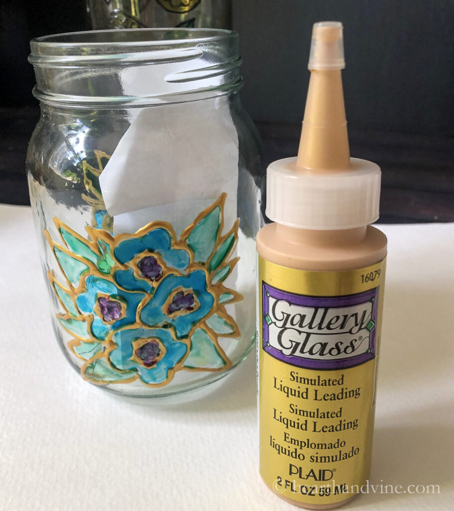 Mason jar with glass paint and gold outline next to a bottle of gold liquid leading.