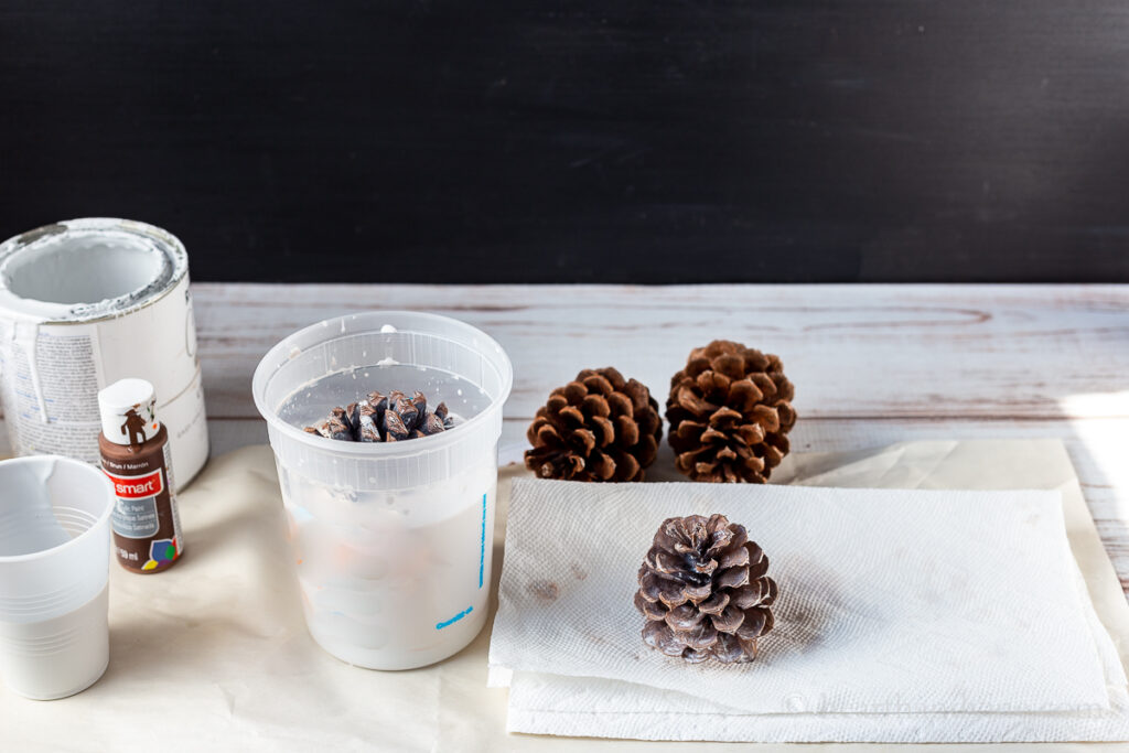 Large plastic container with water and white paint and a pinecone inside. Pinecones and paper towels on the table.