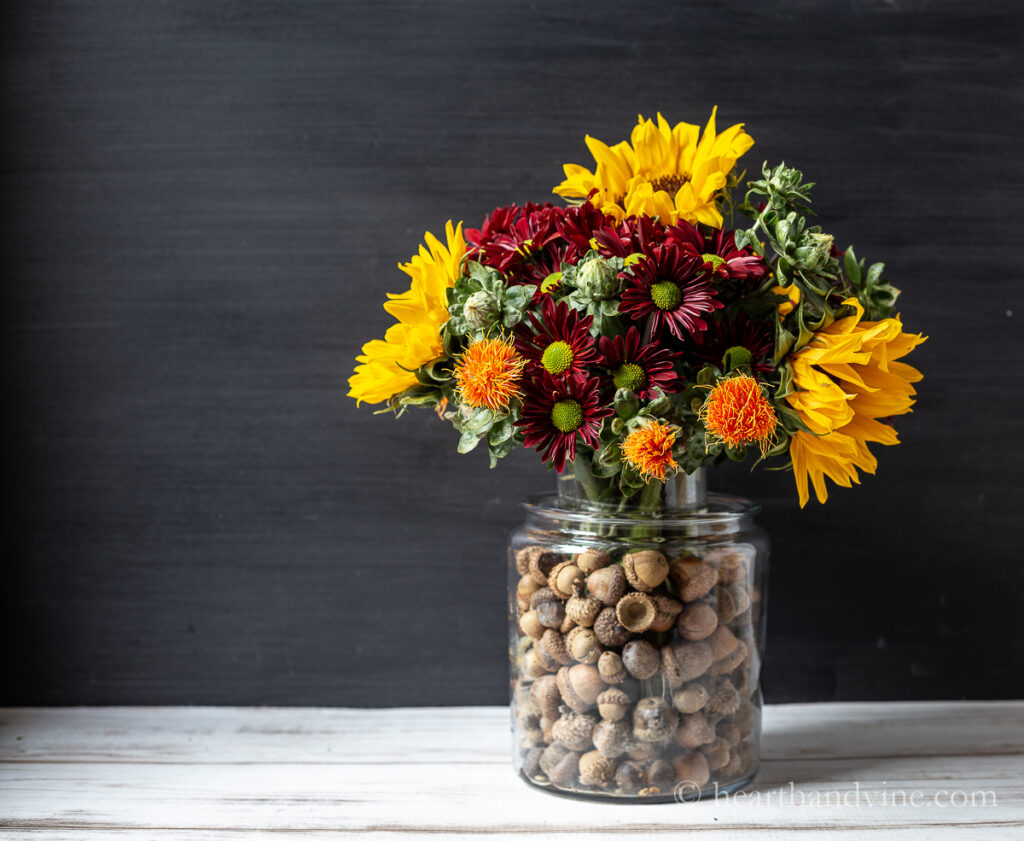 An vase with acorns surrounding another vase with fall flowers.