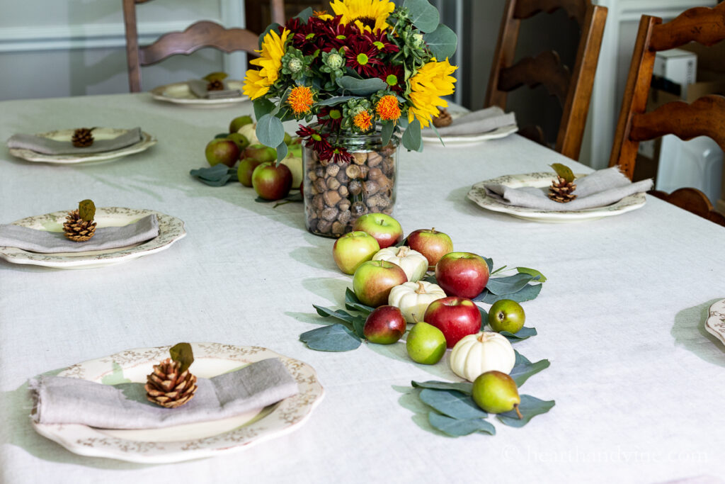 A dining table decorated for fall with gold leaf place cards in pinecones, a fruit and eucalyptus runner and a floral centerpiece with acorns in the vase.
