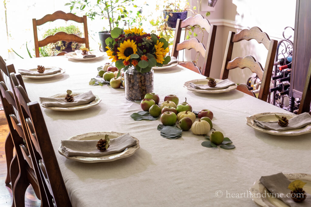 Thanksgiving table decorated with natural materials such as a runner of apples, pears, white mini pumpkins and eucalyptus leaves and a centerpiece vase with acorns and fall flowers.