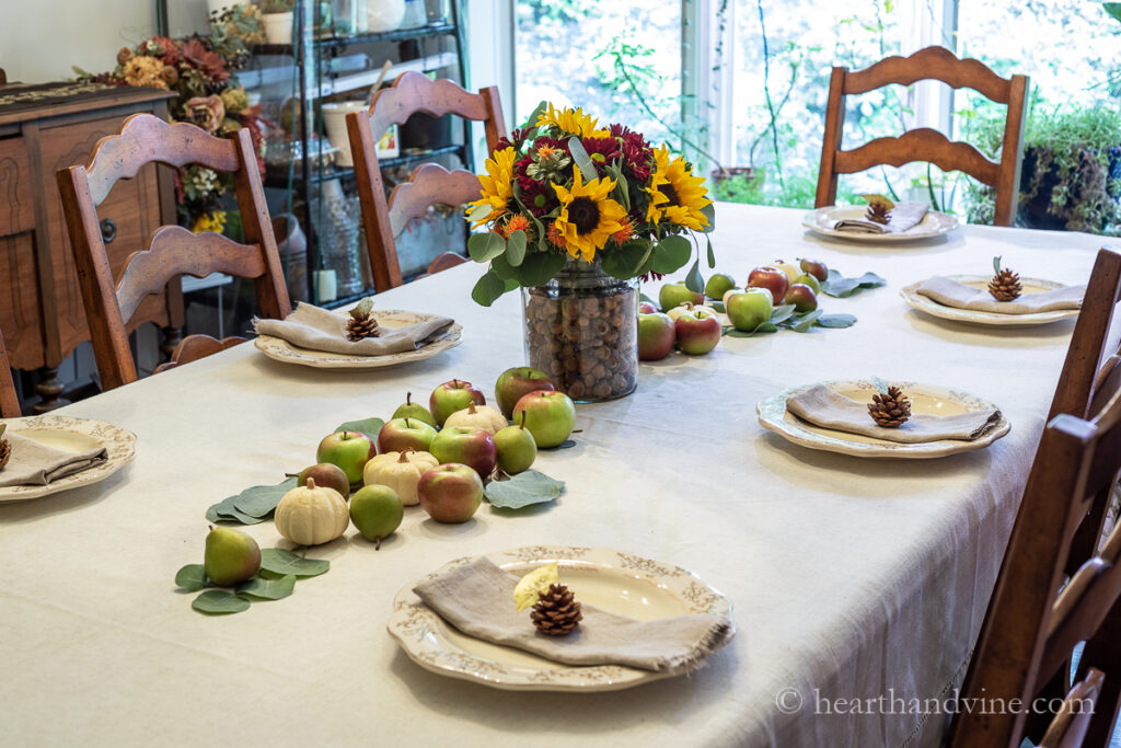 Fall table decorated with an acorn and fall floral centerpiece vase, a runner of apples, mini pumpkins, pears and eucalyptus leaves.