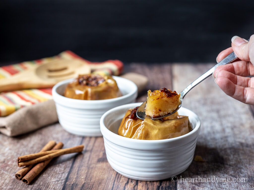 A spoon taking a small section of a baked apple from a ramekin. Another ramekin with a cinnamon baked apple sits behind.