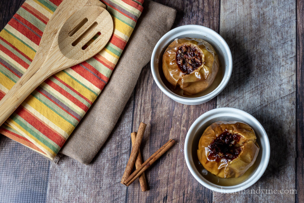 Baked apples in ramekins next to cinnamon sticks, wooden spoons and linens.