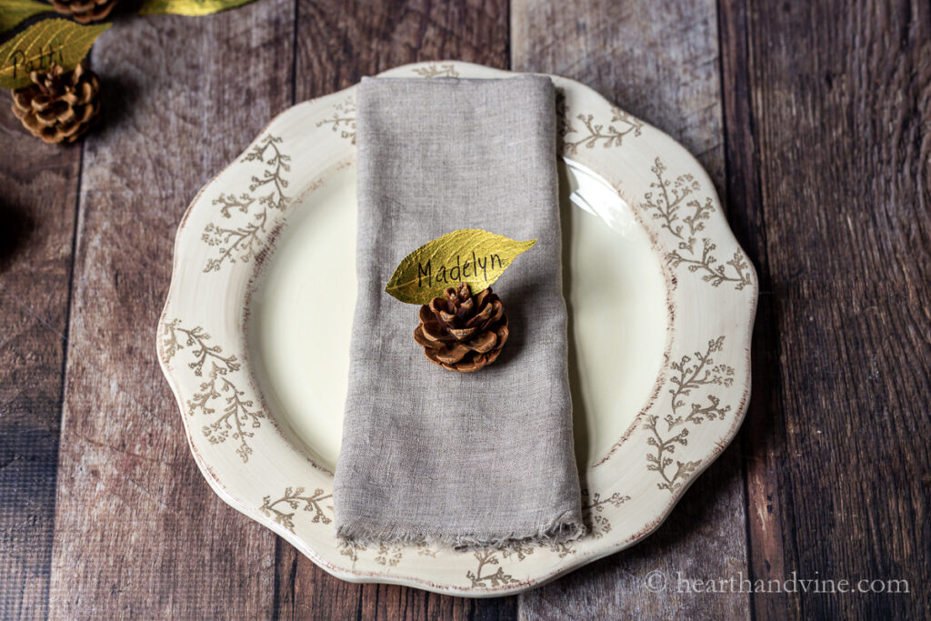 Plate with linen napkin folded in the middle with a pine cone on top holding a leaf place card.