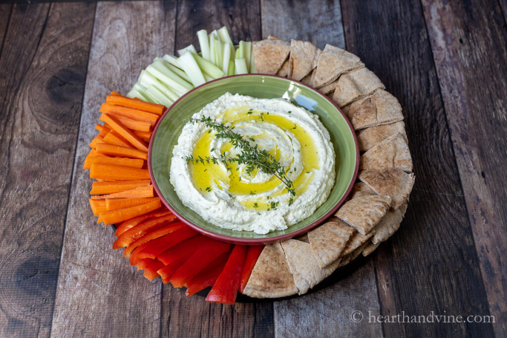 Platter with whipped feta dip in a bowl surrounded by cut up carrots, celery, peppers and pita wedges.