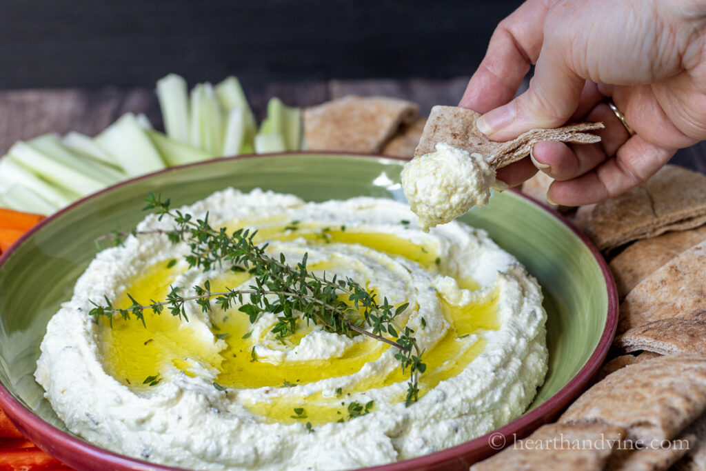 A hand dipping a pita wedge into a creamy cheese dip of feta and cream cheese.