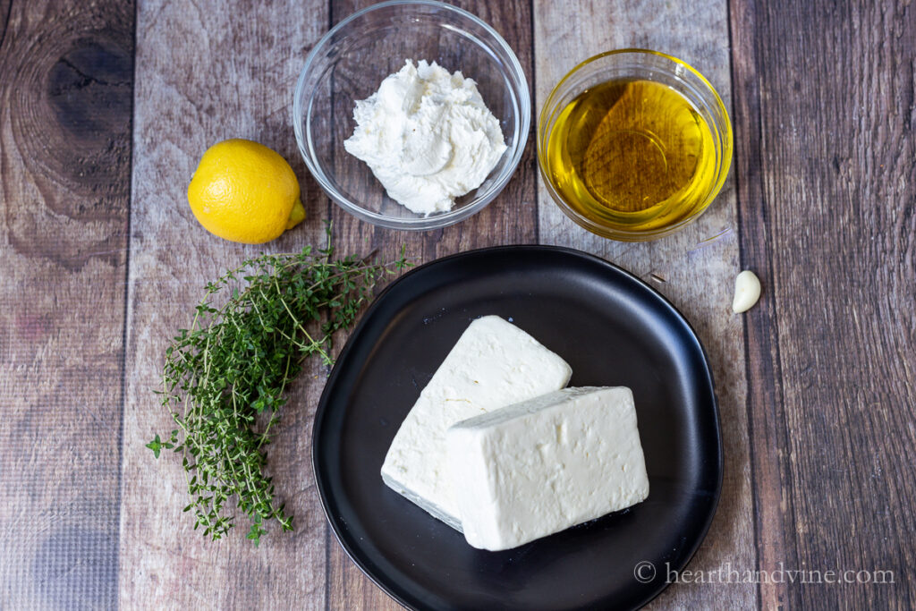Blocks of feta cheese, fresh thyme, lemon, cream cheese, garlic clove and olive oil on a table.