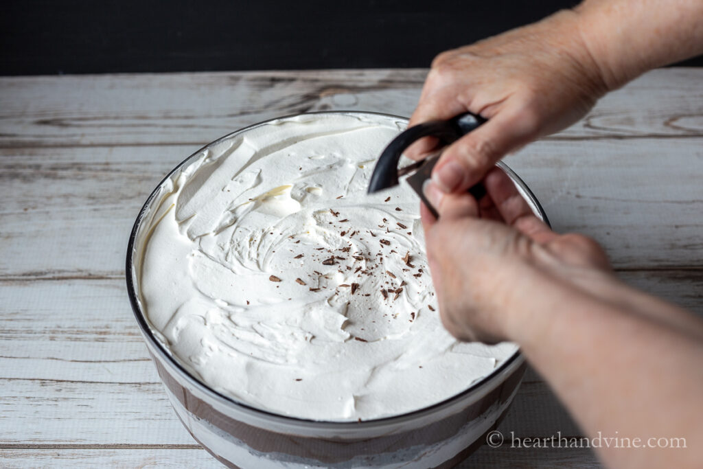 Hands using a vegetable peeler to shave chocolate pieces on top of trifle.