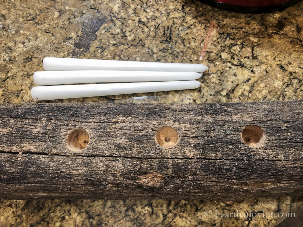 Three white taper candles next to a log with three holes drilled into the top.