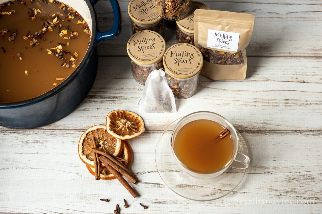 Aerial view of a pot of mulled cider, a glass mug of cider with a cinnamon stick and dried orange slices, cinnamon sticks and clove and jars of spice mix.