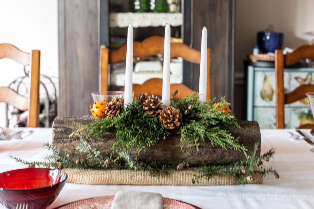 A yule log centerpiece with clove studded clementines, white candles, evergreens and pine cones on a dining table set for Christmas.
