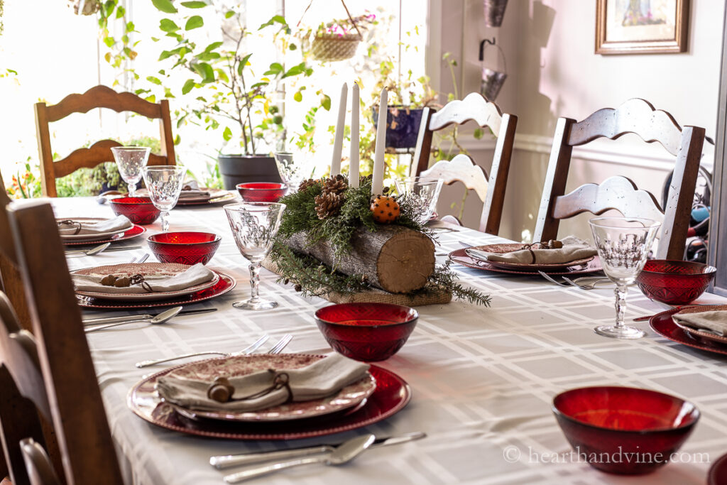 Red and white table settings with a yule log centerpiece decorated with evergreens, pine cones, candles and pomanders.