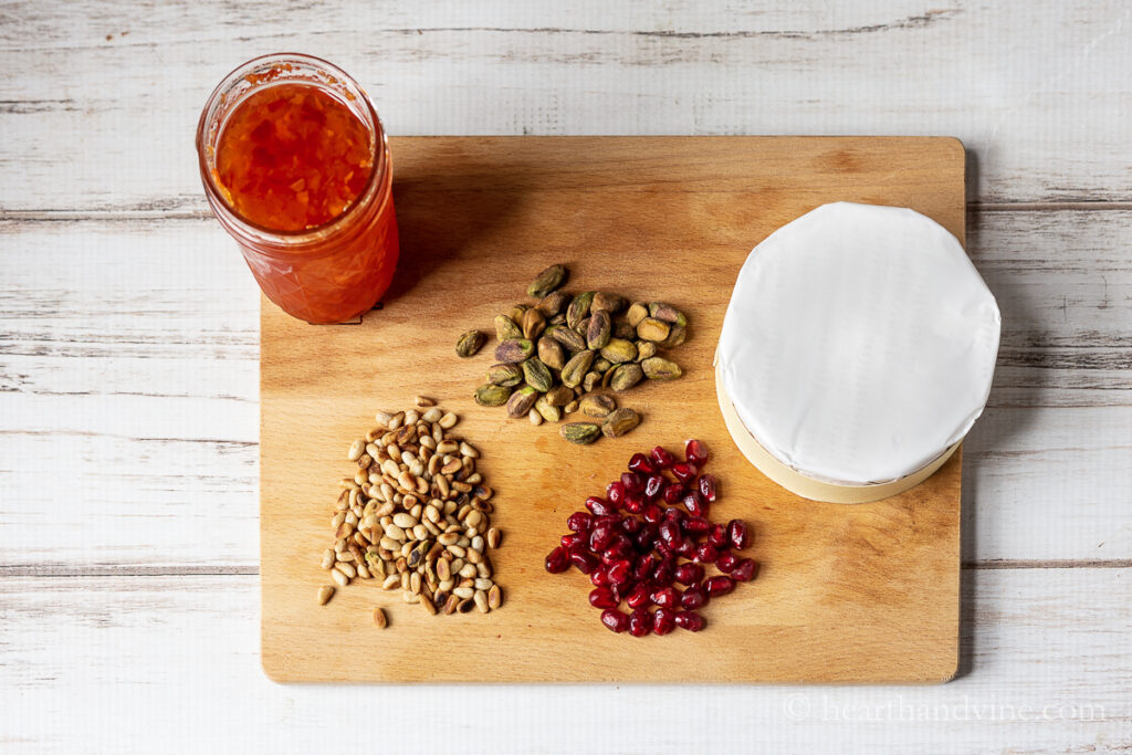 Habanera jam jar, pistachios, roasted pine nuts, pomegranate seeds and a wheel of brie on a wooden board. 