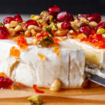 Small knife cutting into a wheel of brie with jam, pine nuts, pistachios and pomegranate seeds.