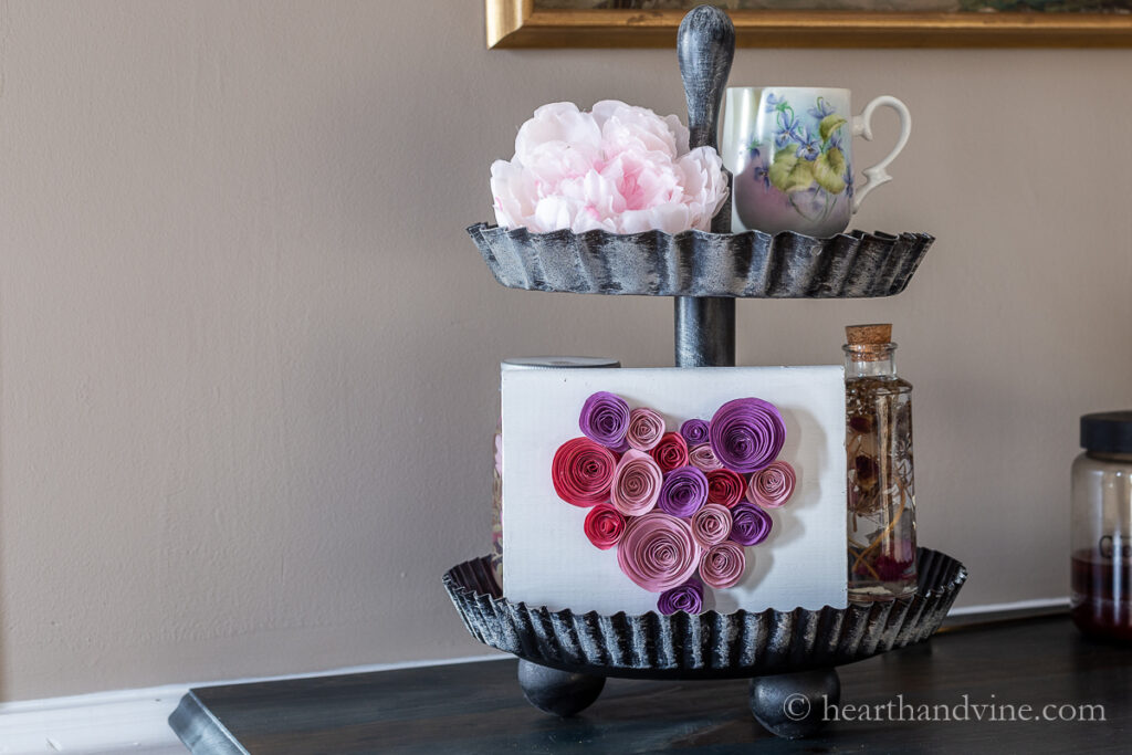 Side view of decorated two tiered tray with wood block with a paper flower heart, faux peony, floral cup and herbarium bottle.
