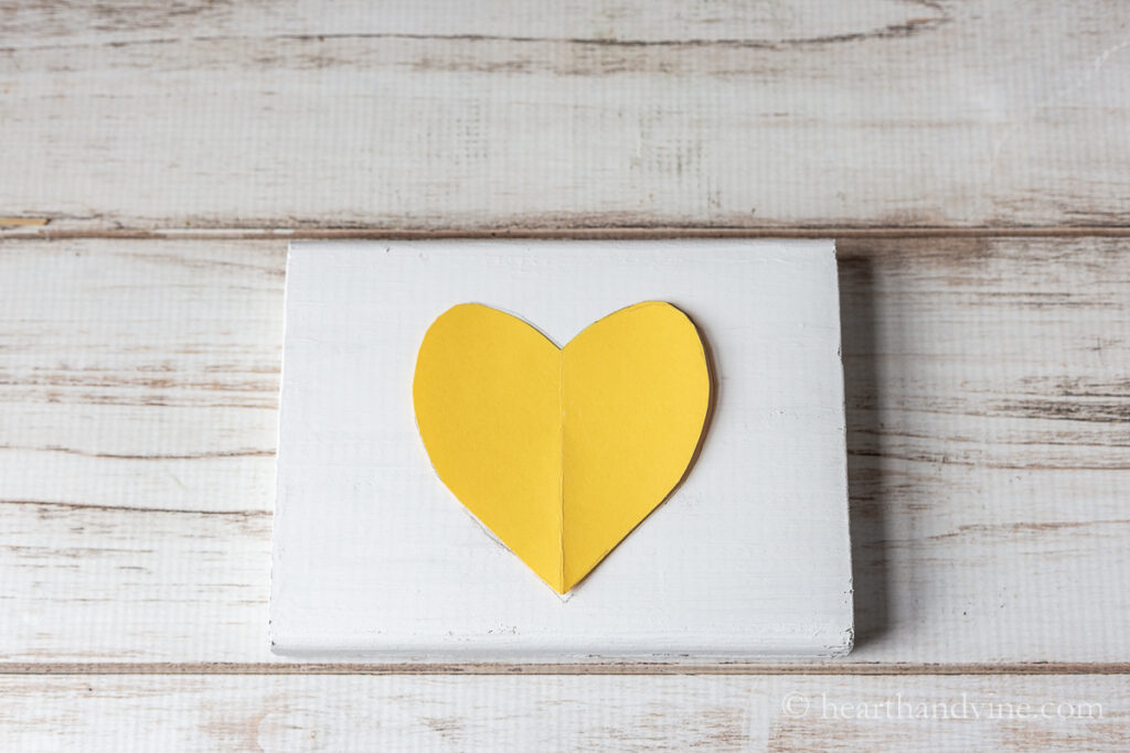 A yellow paper heart outlined in pencil on a white wood block.