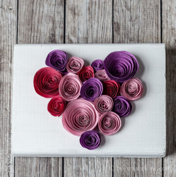 Rolled paper flowers in pinks, red and purple on a wood block in a heart shape.