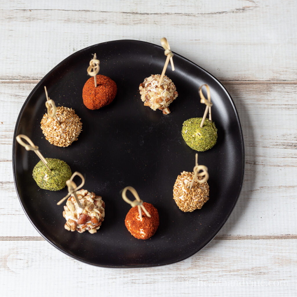 Circle of eight mini cheese balls in different color coating with bamboo toothpicks on a black plate.