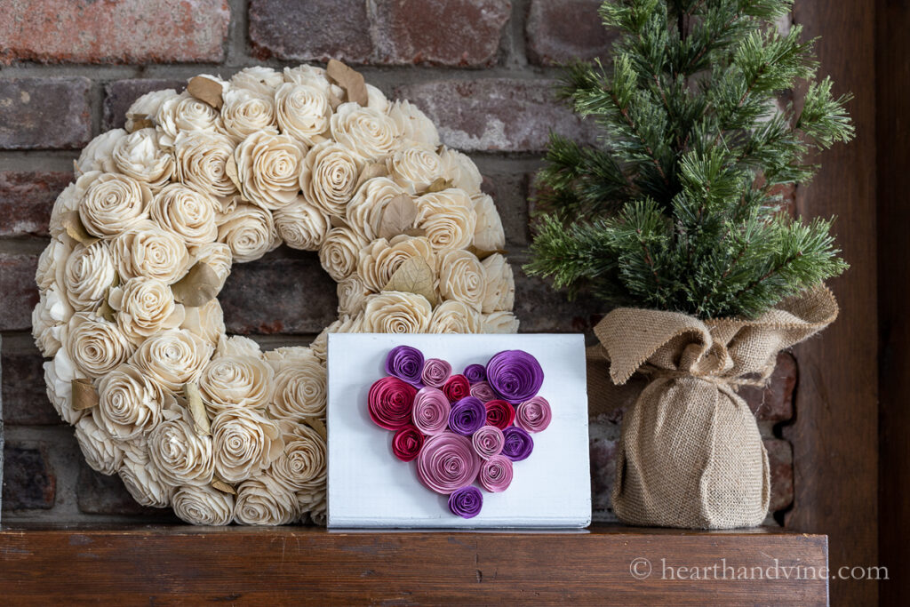 Wooden sign painted white with pink, red and purple paper flowers in the shape of a heart.