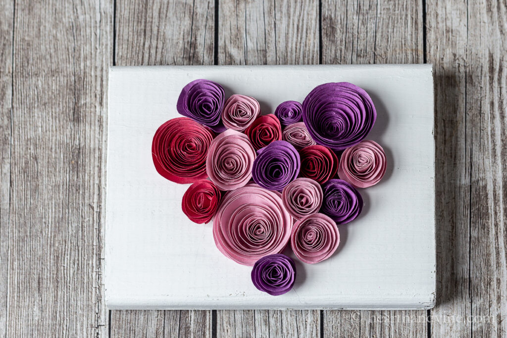 White painted block of wood with a heart shaped rolled paper flower grouping in shades of pink, red and purple.