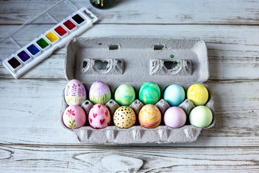A carton of watercolor Easter eggs decorated in different techniques next to a set of kids watercolors.
