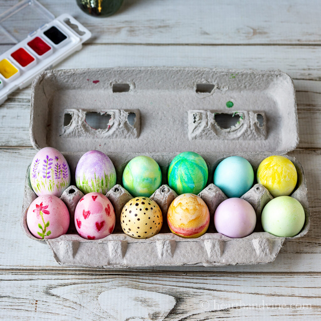 Cardboard carton of egg decorated for Easter with a set of watercolors set on the side.