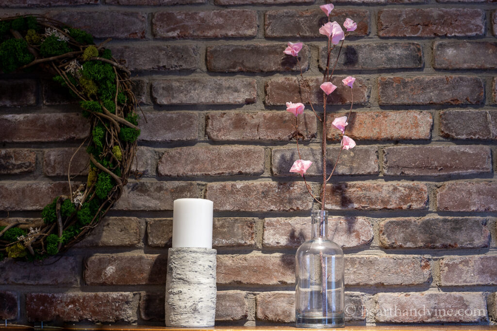 Paper cherry blossoms on a branch in a glass bottle vase on a mantel.