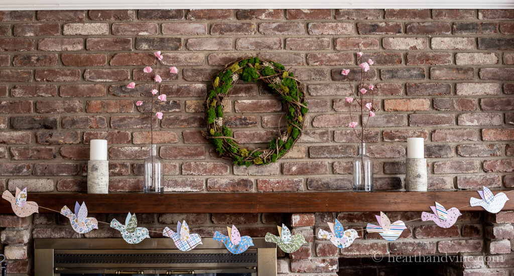 Decorated mantel for spring with a moss wreath, paper cherry blossoms and a paper bird garland.