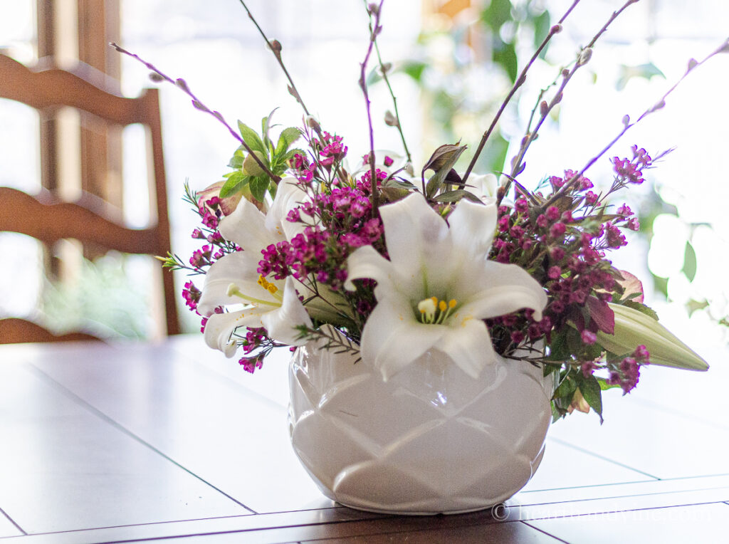 Flower arrangement in a white bowl with Easter lily flowers, pussy willow stems, pink wax flower and helebores on a dining room table.