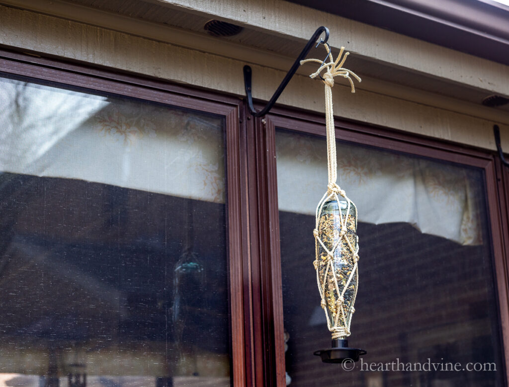 A beautiful macrame hanging bird feeder on the side of a porch.