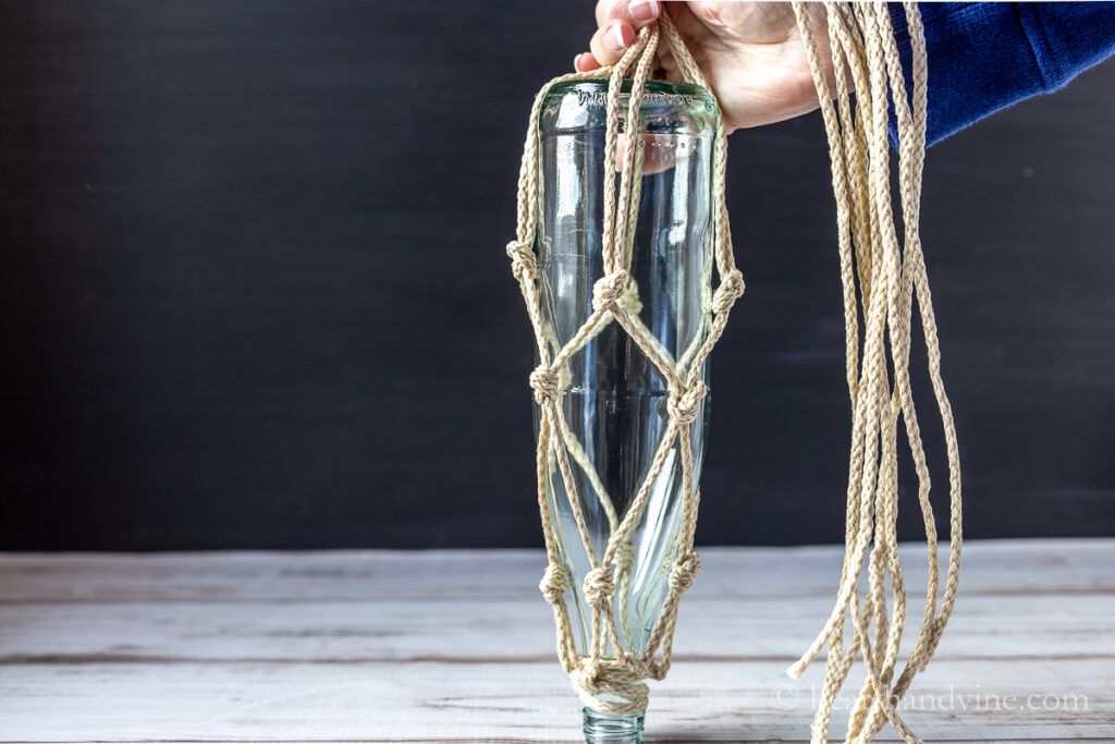 Macrame hanging on a bottle upside down with a hand holding remaining untied cords.