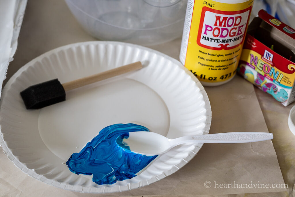 A paper plate with a mixture of Mod Podge and neon food coloring next to a sponge brush.